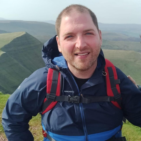 Andrew is standing on the top of a mountain. He is wearing a blue jacket and a rucksack.